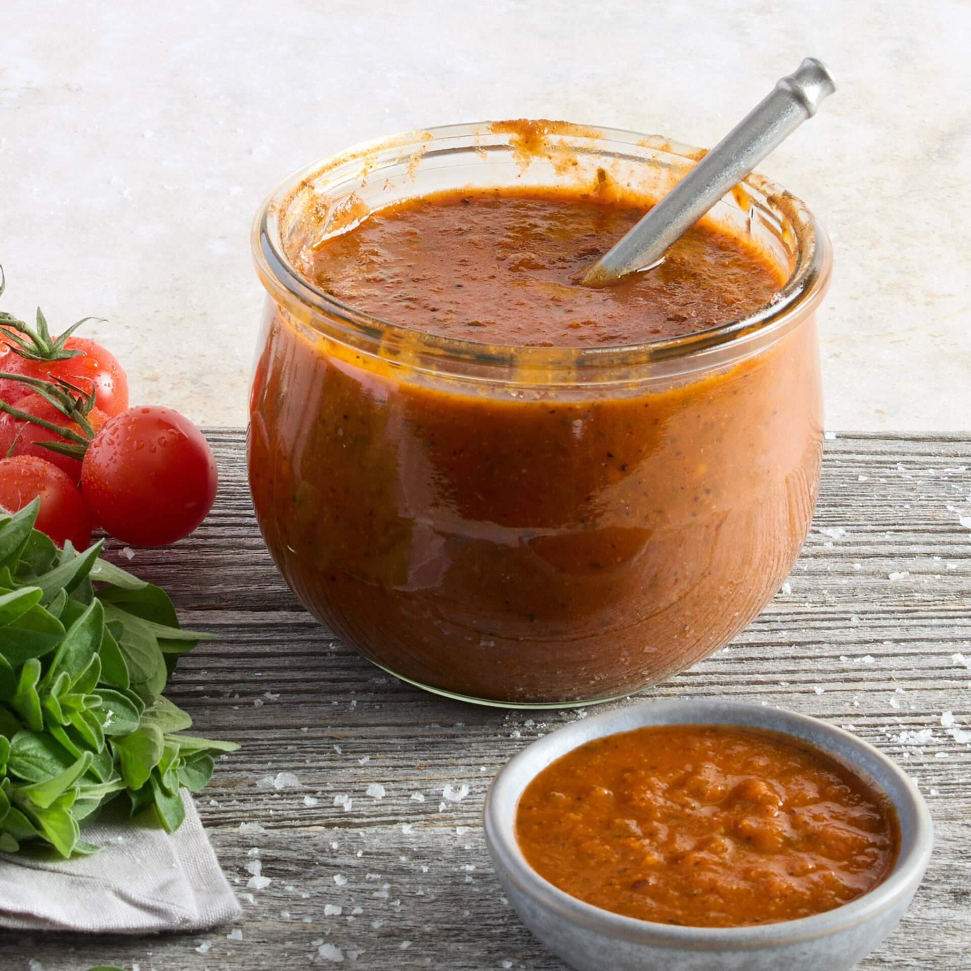 Jar of Smoky Chipotle BBQ sauce presented next to a side dish of sauce next to a fresh bunch of herbs and cherry tomatoes.