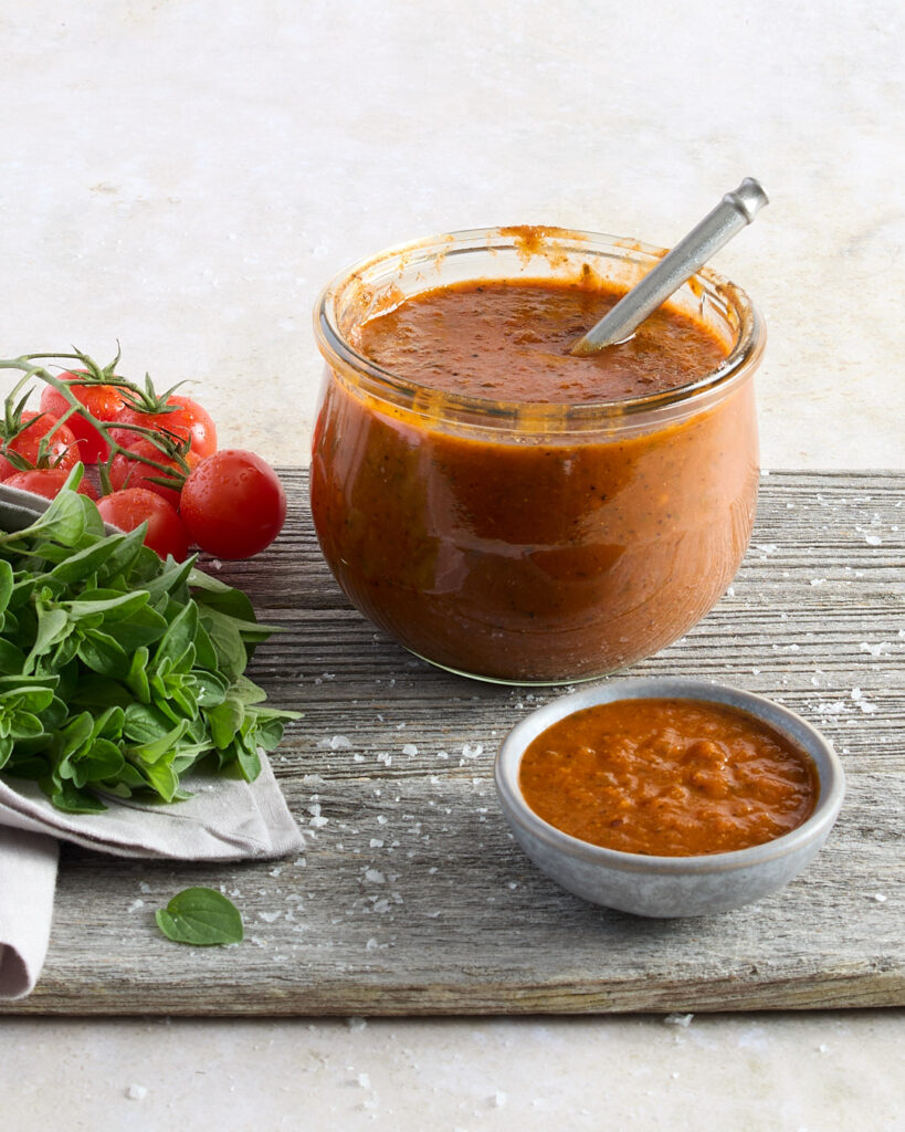 Jar of Smoky Chipotle BBQ sauce presented next to a side dish of sauce next to a fresh bunch of herbs and cherry tomatoes.