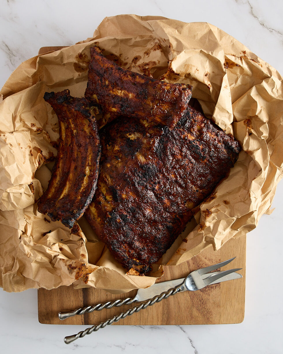 Caramelised barbecued ribs sitting on the brown paper bag on a cutting board with serving utensils.