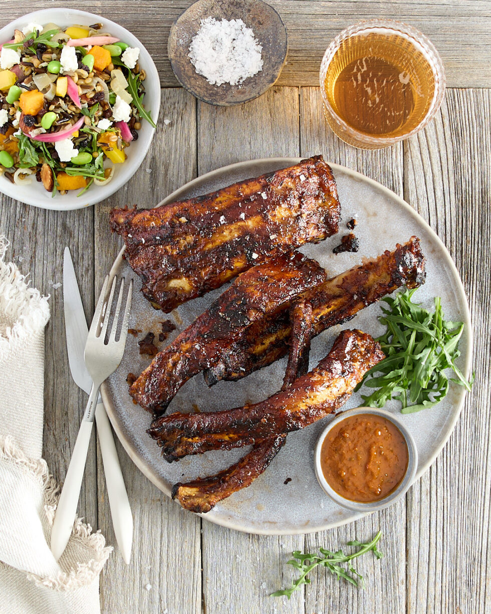 A rustic plate of ribs served with a side dish of smoky chipotle BBQ sauce and kitchen sink pumpkin salad.