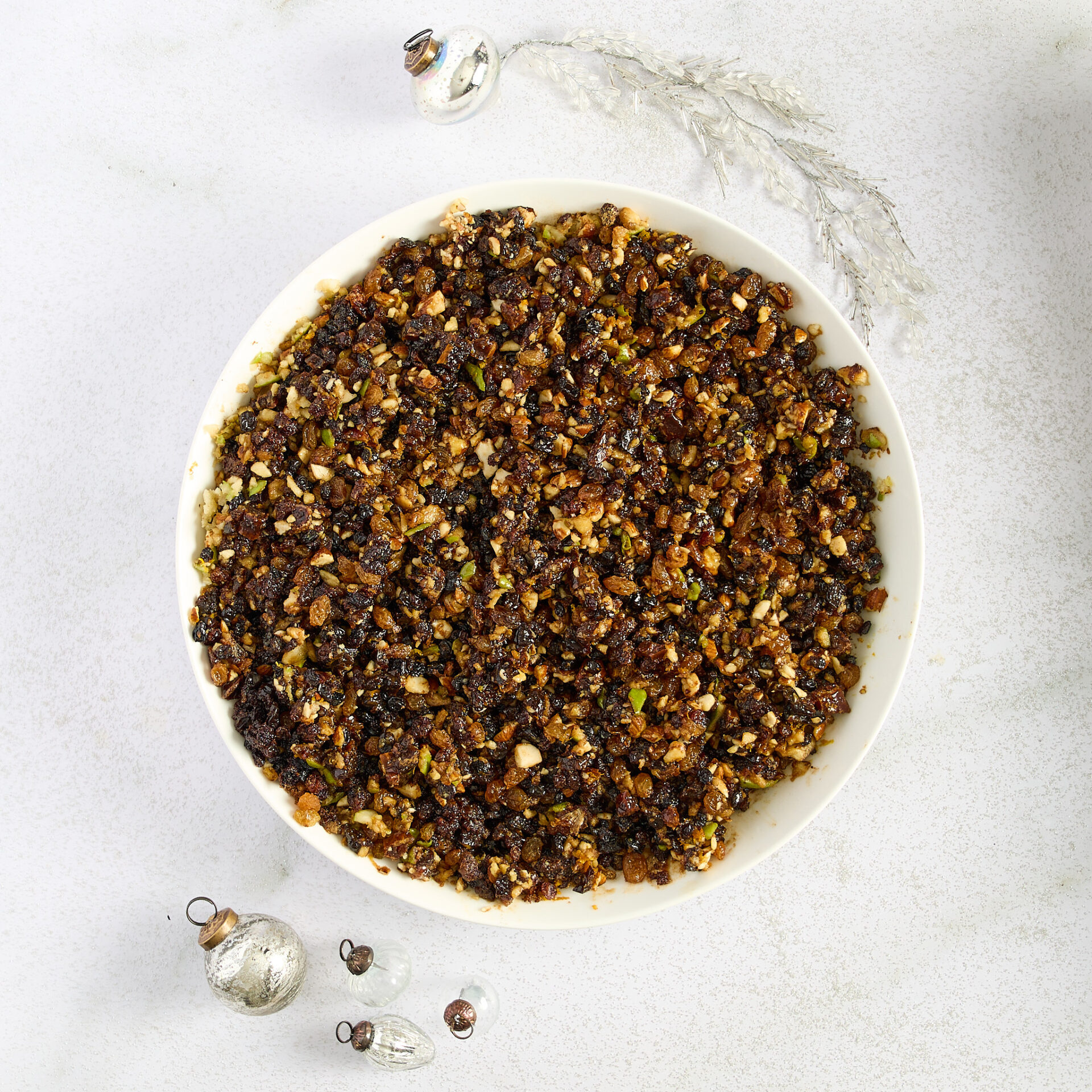 On a white marble bench, a white ceramic bowl holds homemade fruit mince, surrounded by Christmas decorations.