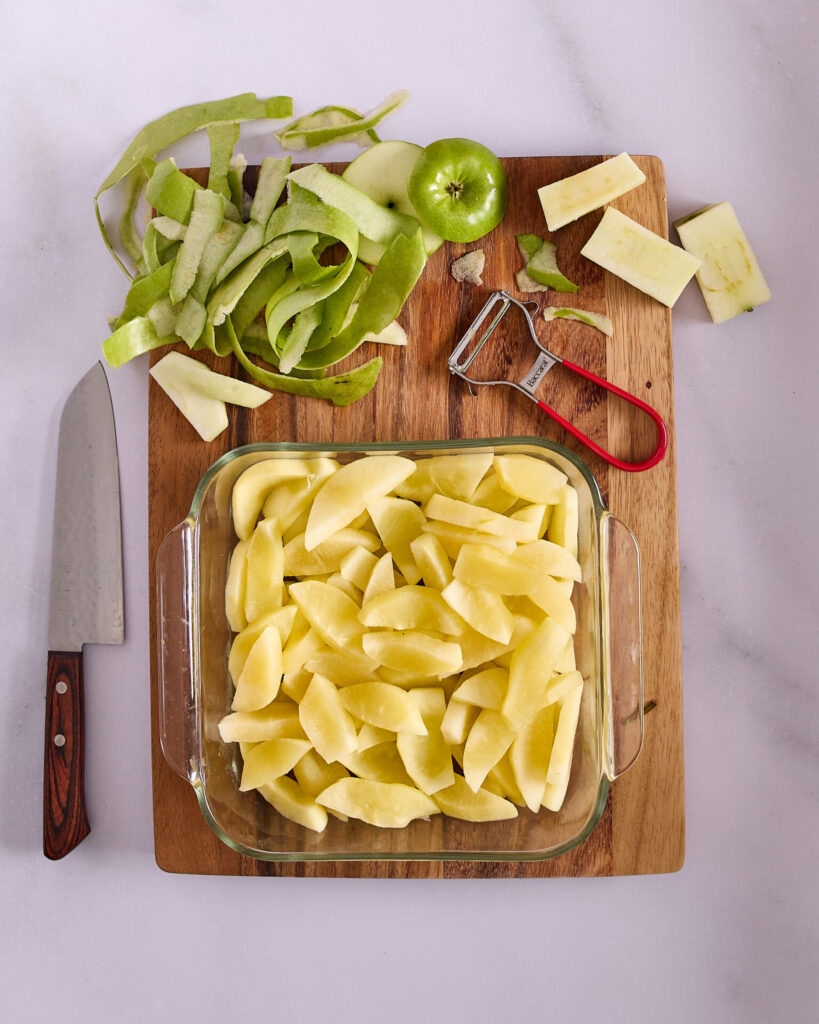 Pre-cooked granny smith apple slices in a square glass dish on a cutting board still cluttered with apple peels and cores.