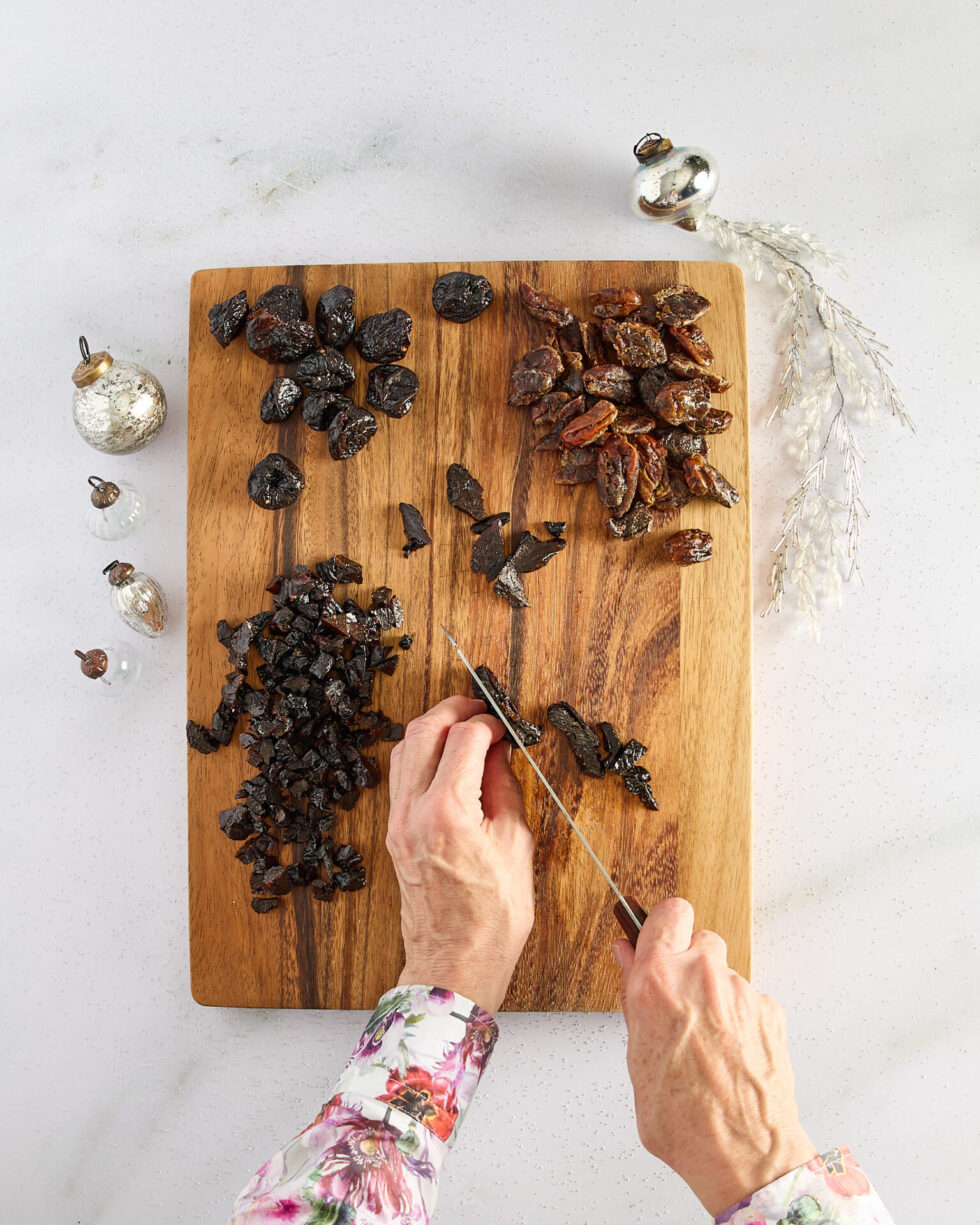 Roughly chopping larger dried fruits on a wooden cutting board with festive Christmas decorations about.