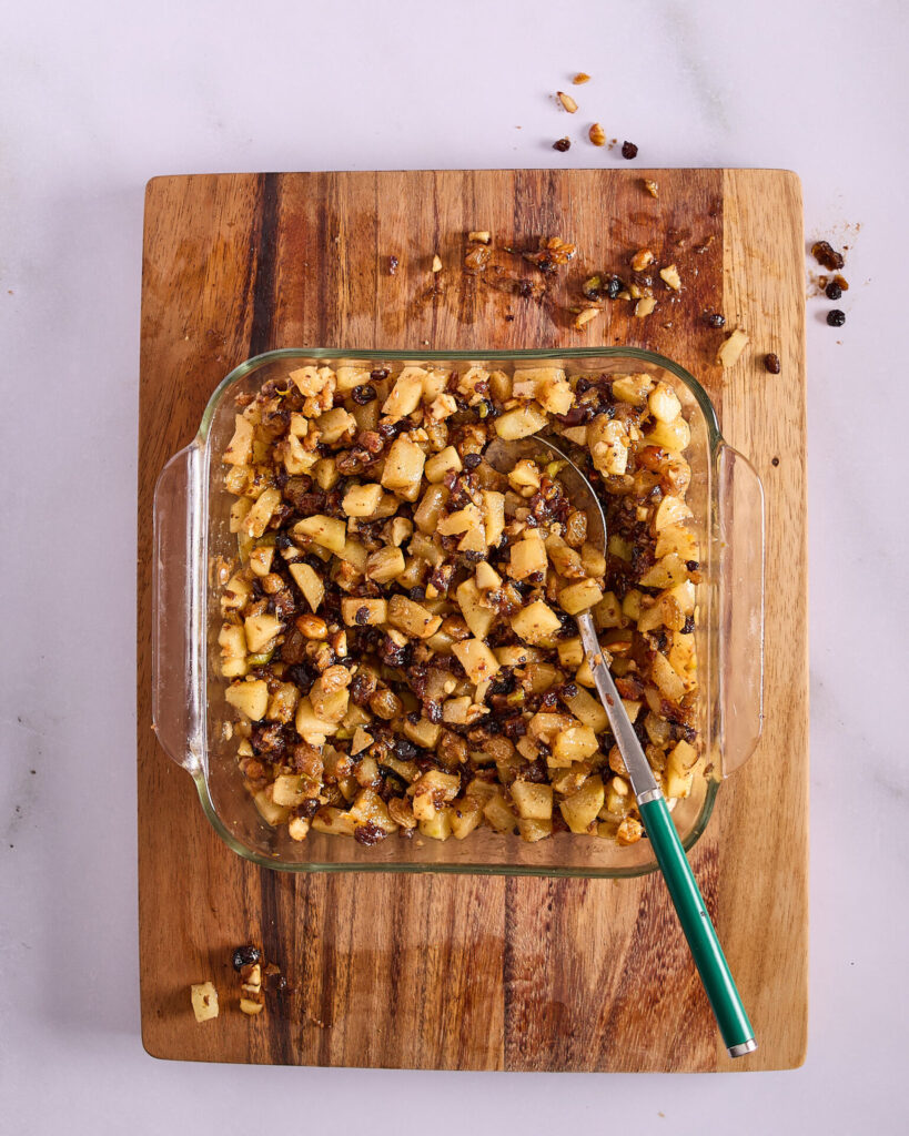 Chopped apples stirred into a glass dish of fruit mince sitting on a wooden cutting board.