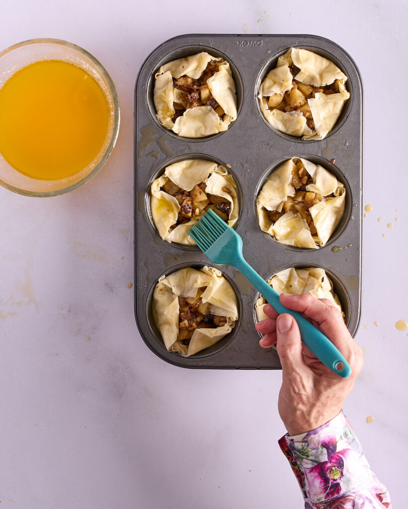 Cook is brushing six apple fruit mince tarts in their tin with melted butter before baking.