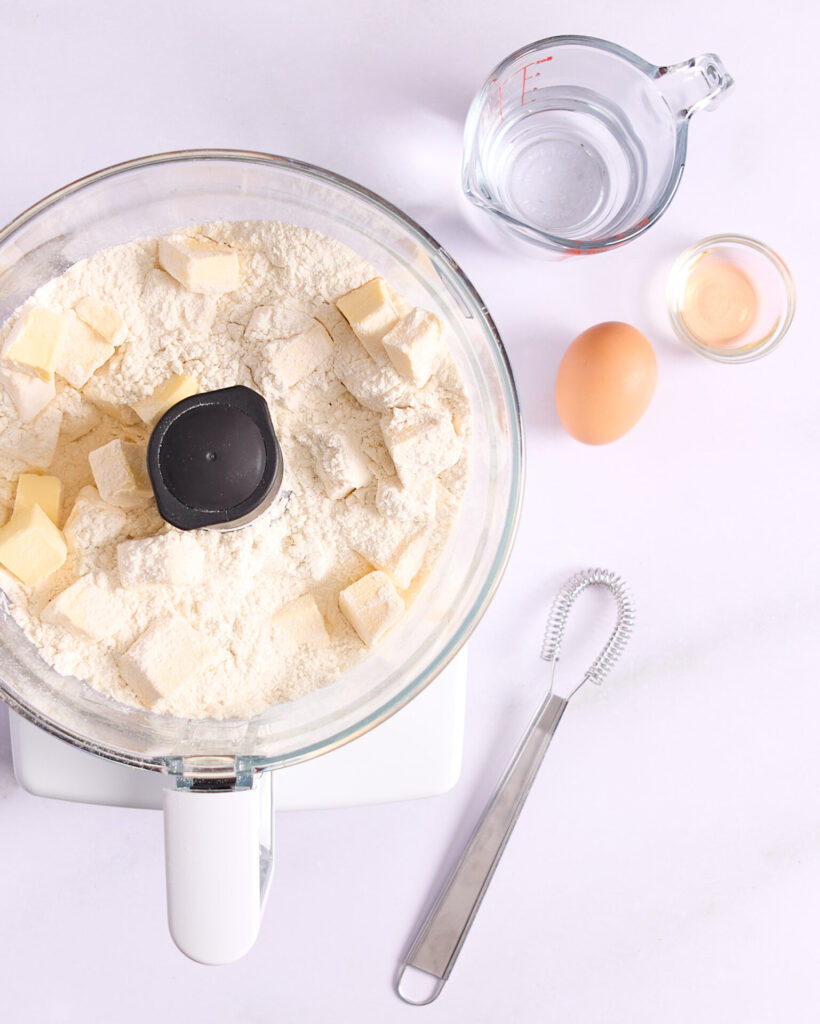 Dry ingredients in a food processor bowl with frozen cubes of butter ready for blending.