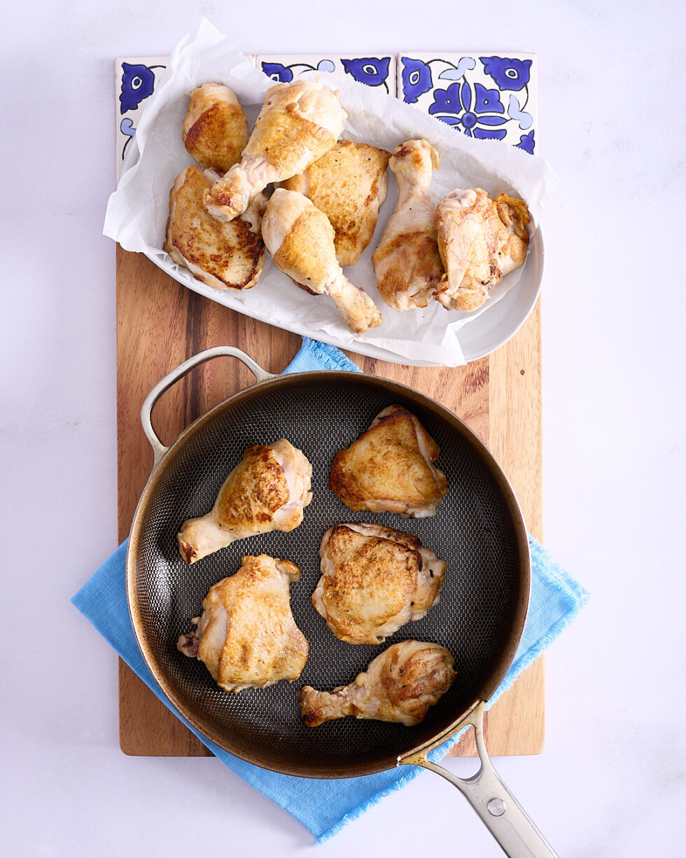 Browned chicken pieces in batches with some in a frying pan with some draining in a white ceramic dish lined with paper towel on a wooden cutting board.