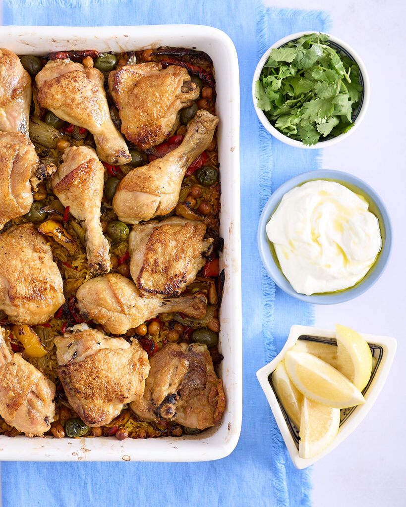Moroccan chicken bake ready to serve from the oven with bowls of coriander, Greek yoghurt condiment and lemon wedges on a pale blue napkin.