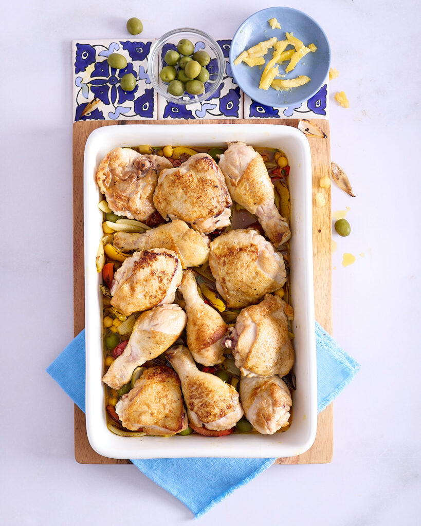 Chicken pieces has been layered over the flavour base in a white ceramic baking dish on a wooden cutting board.