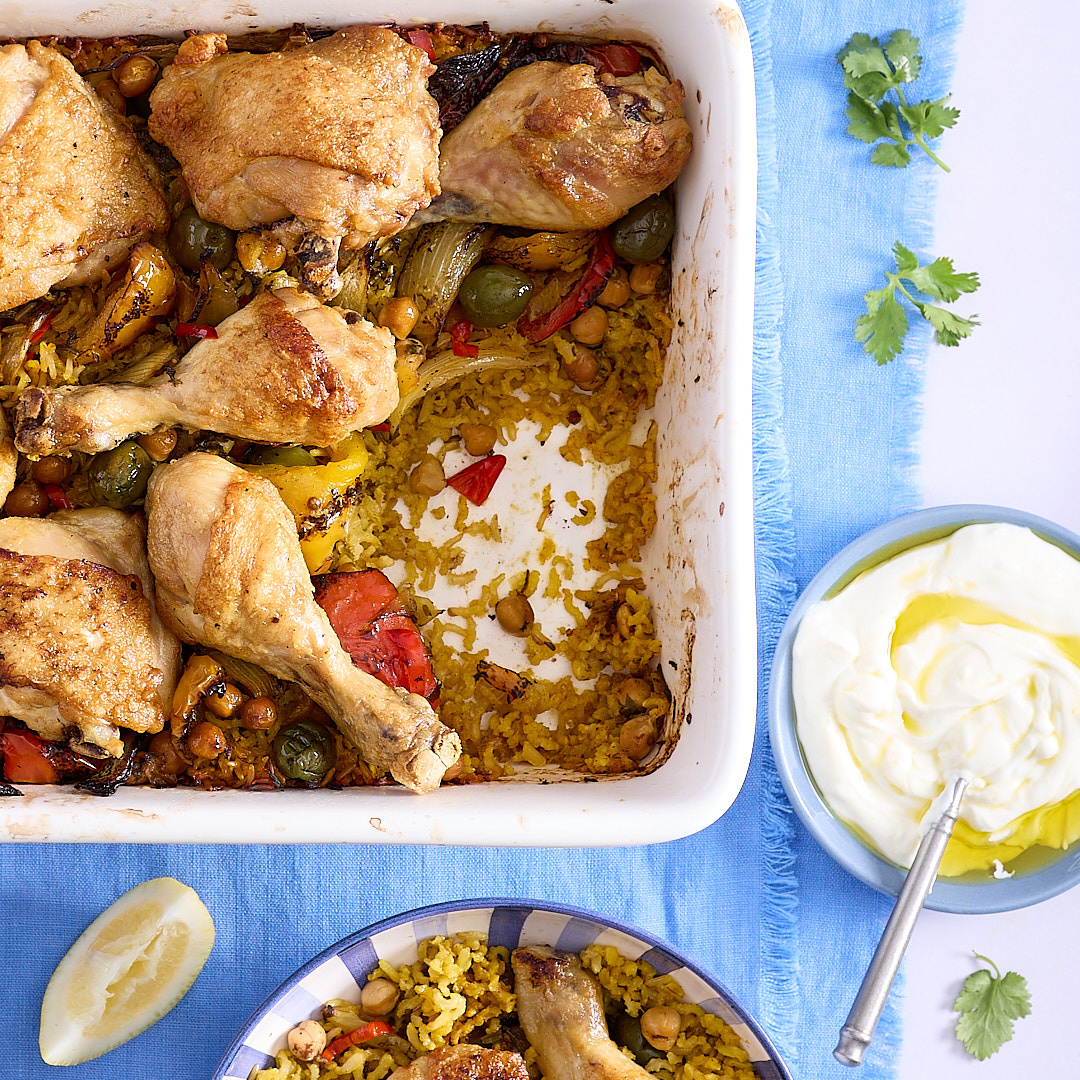 One pan Moroccan chicken and rice in a white ceramic baking dish, a serving ready to eat, and a bowl of the Greek yoghurt condiment on a blue linen napkin.
