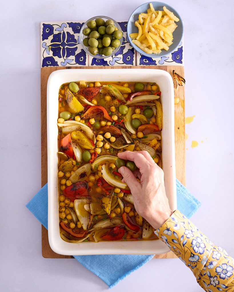 Sandra starts adding the Moroccan flavoured rice base in a white ceramic baking dish on a wooden cutting board with bowls of green olives and preserved lemon.