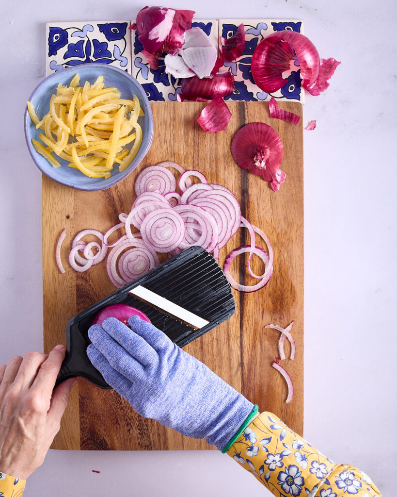 Sandra wearing a cut resistant glove to thinly slice red onion using a mandolin over a wooden cutting board.