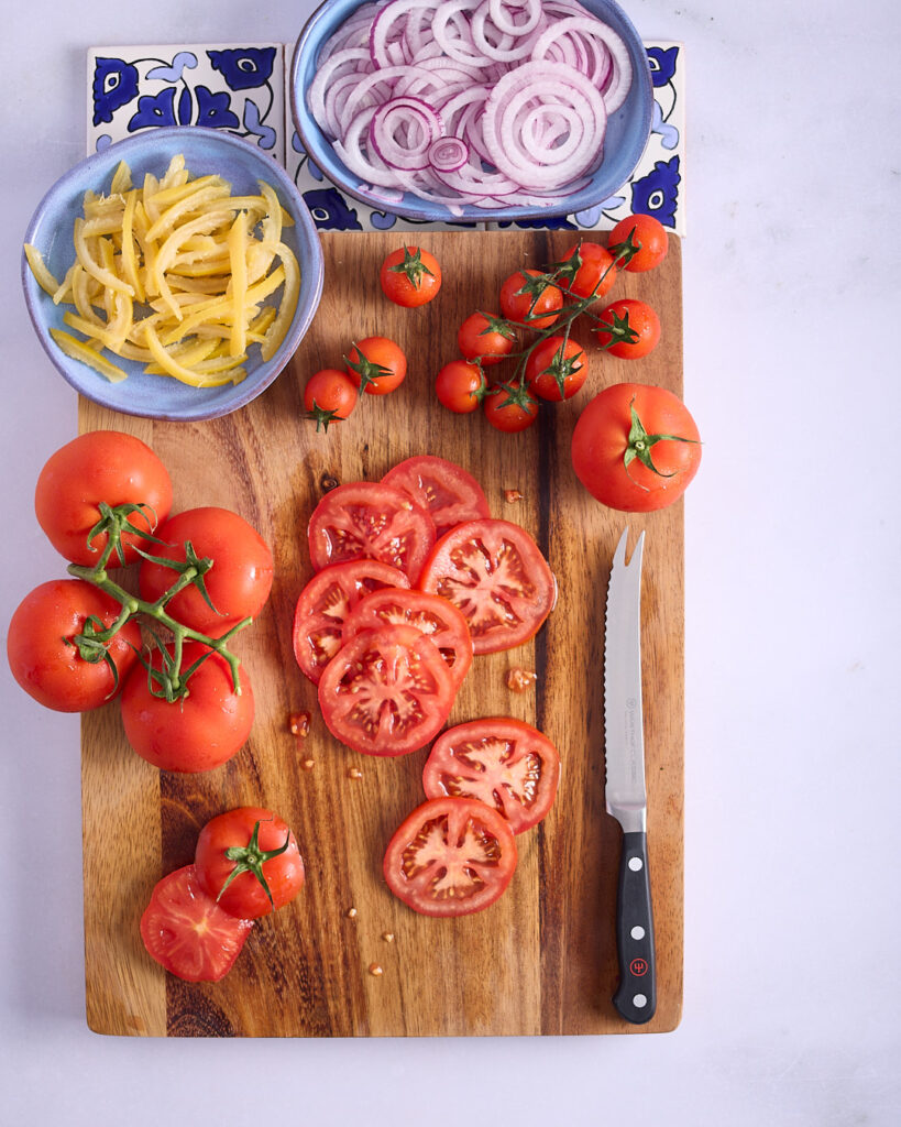 Large and small summer fresh tomatoes sit on a wooden cutting board with dishes of sliced preserved lemon and red onion.