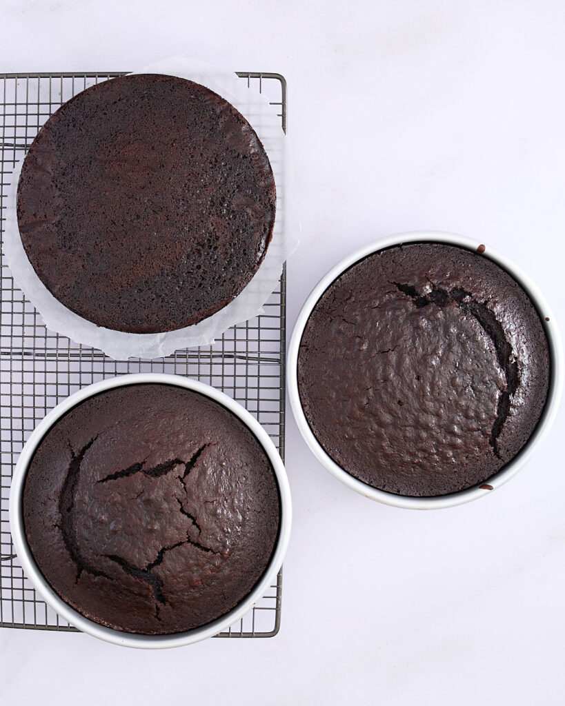 Overhead view of three baked devil’s food cake layers, two still in tins with surface cracks and one inverted on a cooling rack, showing the texture and colour of a rich chocolate layer cake.