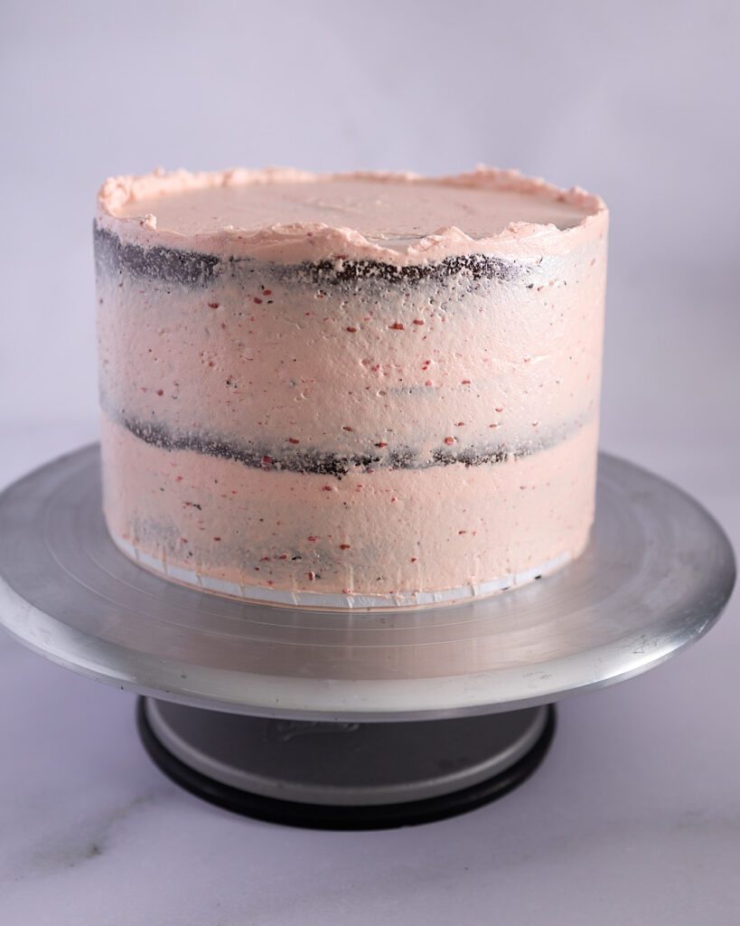 Chocolate layer cake on a turntable with a thin crumb coat of pale pink raspberry buttercream applied, showing visible layers through the frosting.