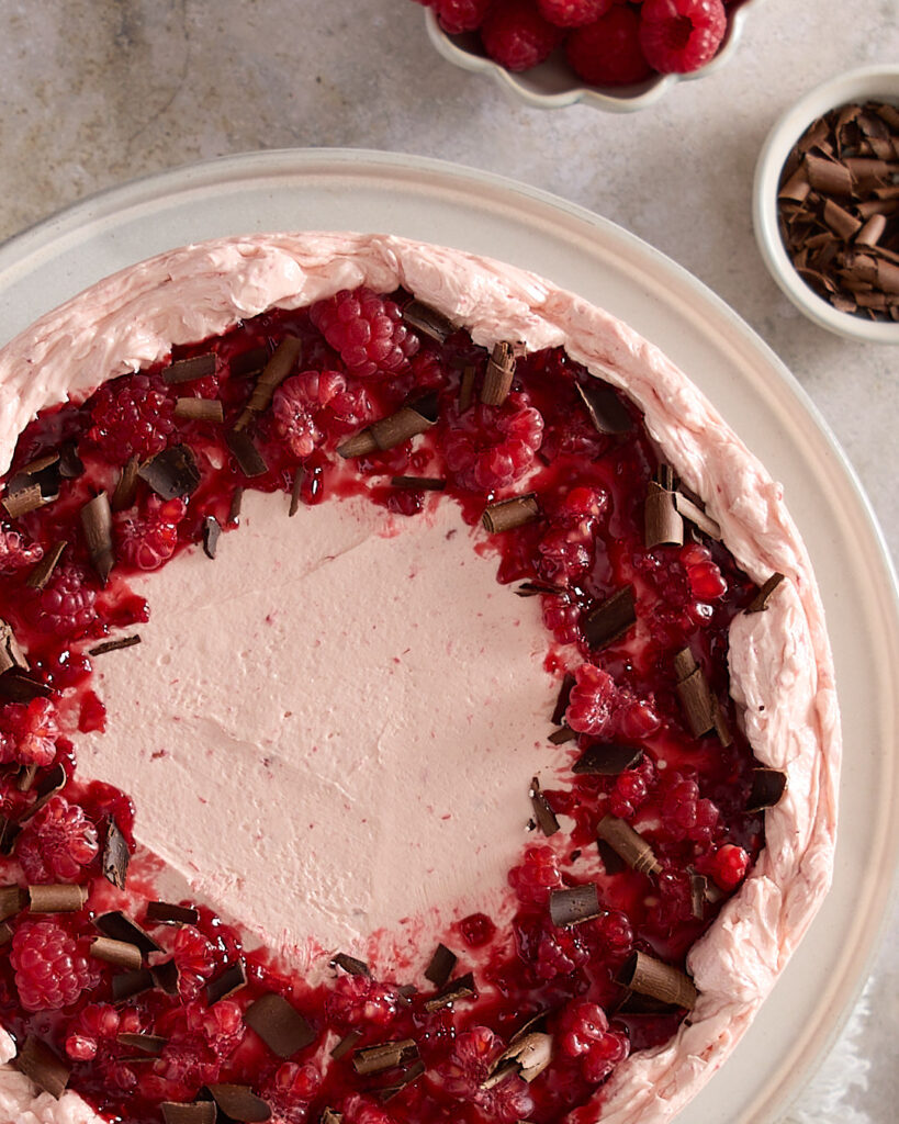 Overhead close-up of the top of a chocolate raspberry celebration cake, decorated with raspberry Swiss meringue buttercream, spooned raspberry jam, fresh raspberries, and dark chocolate curls, arranged in a loose, rustic ring around the edge.