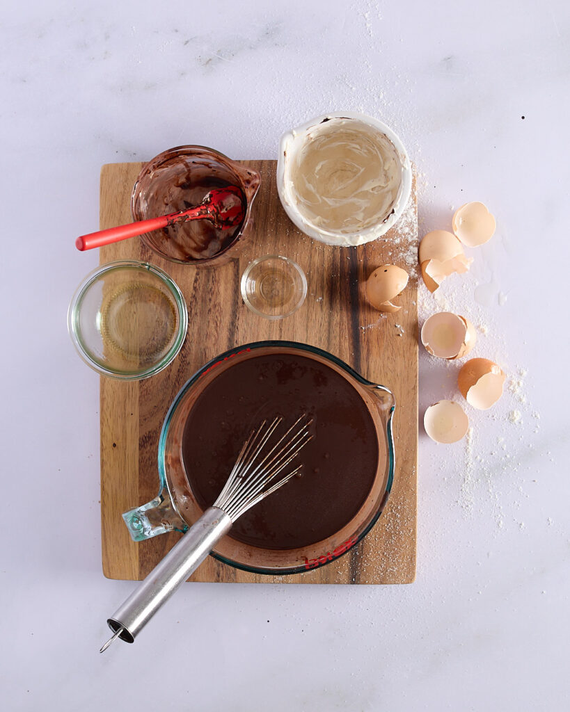 Overhead view of whisked wet ingredients in a glass jug for devil’s food cake, surrounded by empty bowls, eggshells, and measuring cups on a wooden board.