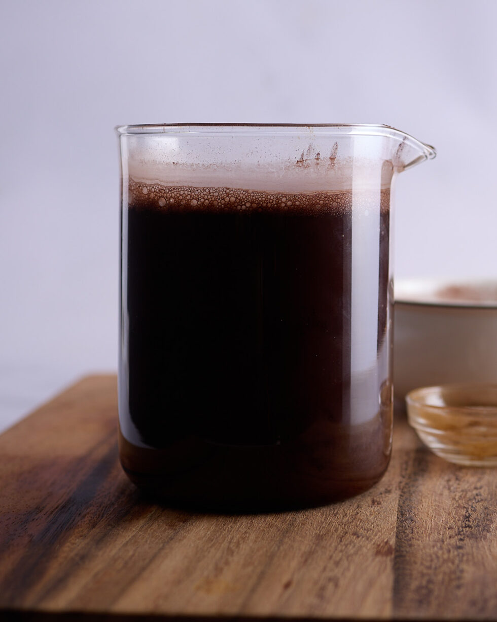 Glass jug filled with bloomed cocoa powder and espresso powder in boiling water, resting on a wooden board as part of a devil’s food cake preparation.