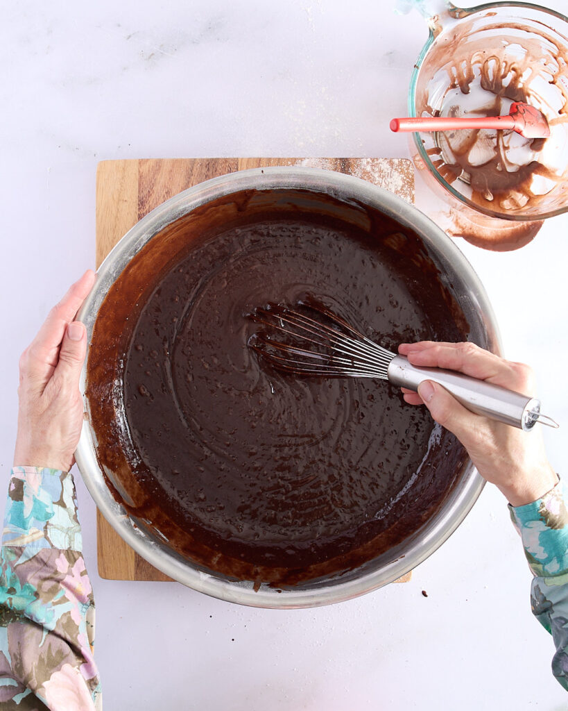 Sandra gently whisking chocolate cake batter in a large mixing bowl, showing the smooth, glossy texture of a fully combined devil’s food cake batter. A jug with leftover mixture sits nearby on a wooden board.