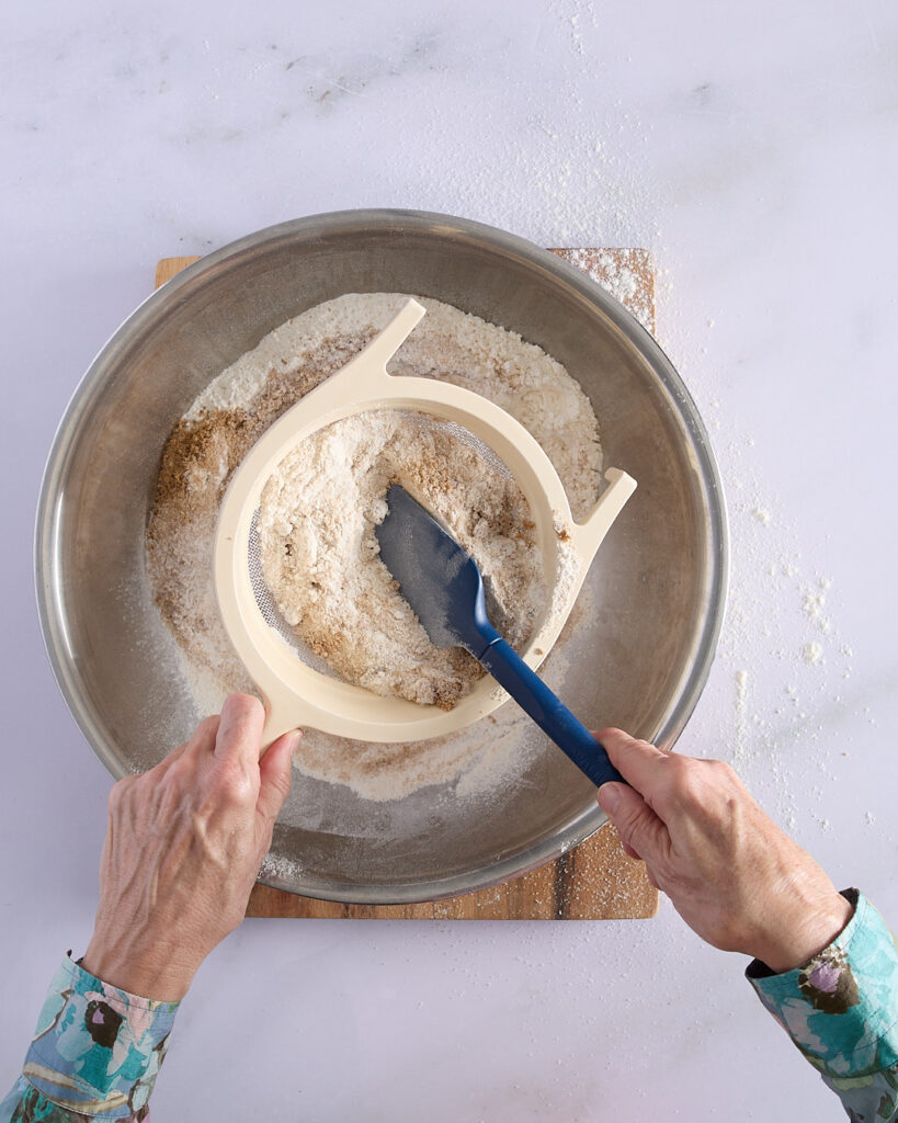 Sandra sifting flour, brown sugar, white sugar, and leavening agents through a coarse sieve into a large metal mixing bowl while preparing a devil’s food cake.