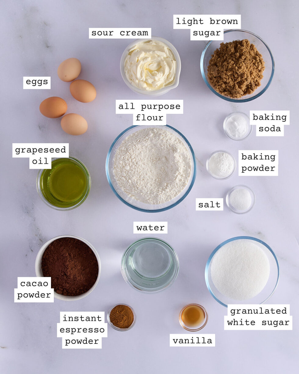 Ingredients for a three-layer devil’s food cake recipe laid out on a marble surface, including flour, cocoa powder, espresso powder, sugars, eggs, sour cream, grapeseed oil, vanilla, and leavening agents.