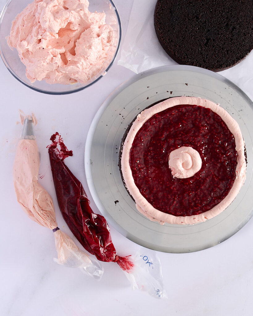 Partially assembled three-layer chocolate cake with raspberry jam and raspberry Swiss meringue buttercream filling piped between layers, shown on a cake turntable with piping bags and a bowl of buttercream.