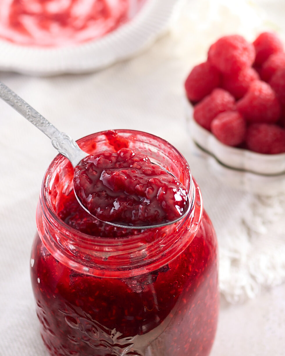 A spoon scooping vibrant raspberry jam from a glass jar, with a small bowl of fresh raspberries and a white dish blurred in the background.