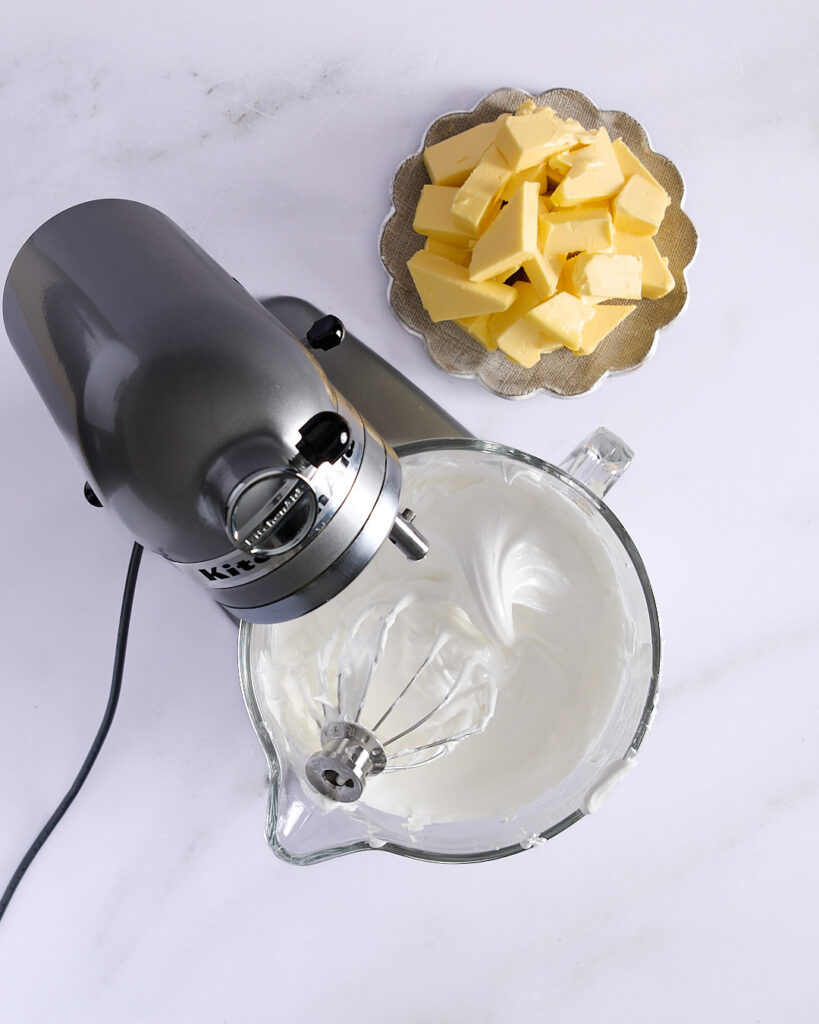 Stand mixer with glossy, stiff-peaked meringue and a plate of cubed unsalted butter beside it, ready for the next step in buttercream preparation.
