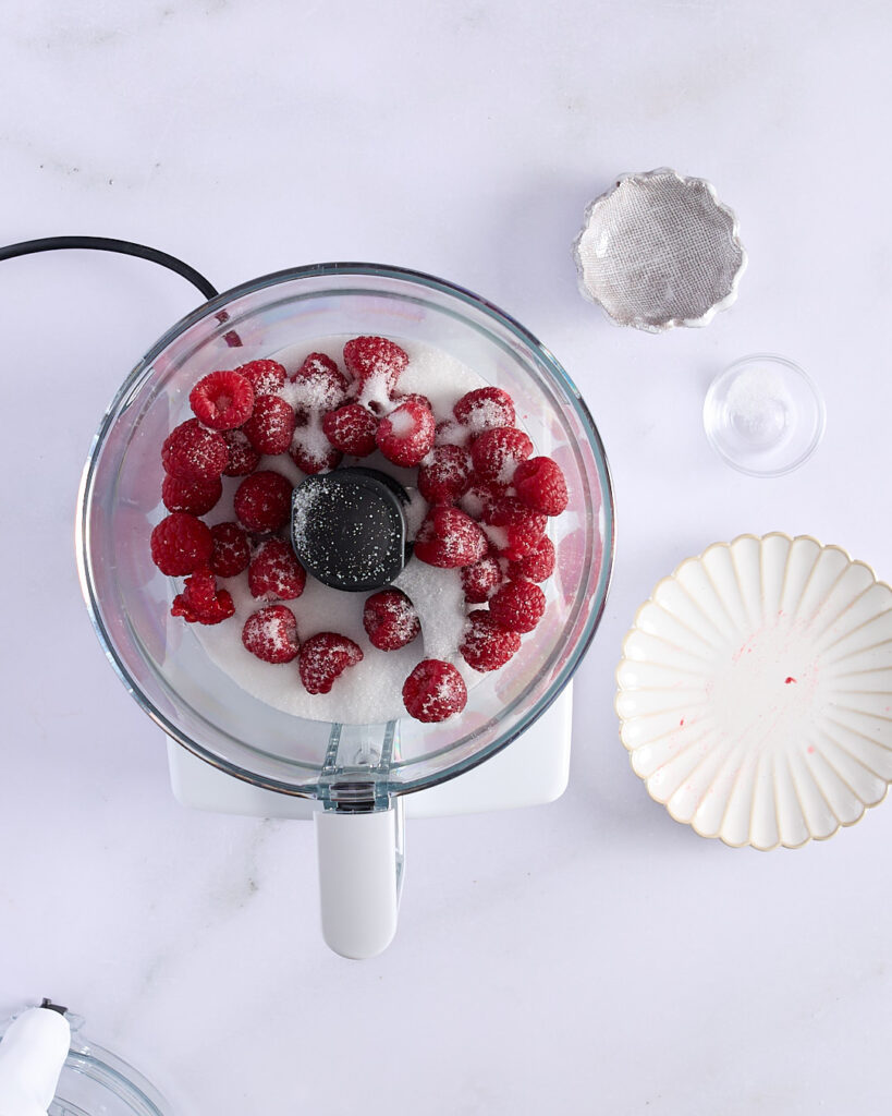 Fresh raspberries, granulated white sugar, and a pinch of salt in a food processor bowl, ready to be blended into raspberry purée for Swiss meringue buttercream.