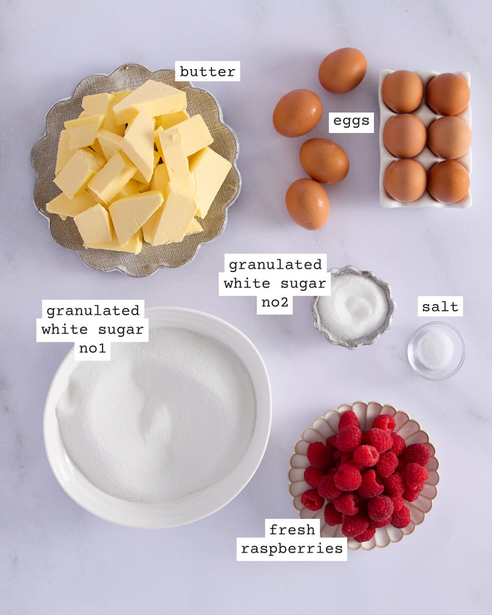 Ingredients for raspberry Swiss meringue buttercream laid out on a marble surface, including eggs, unsalted butter, fresh raspberries, granulated white sugar, and salt.