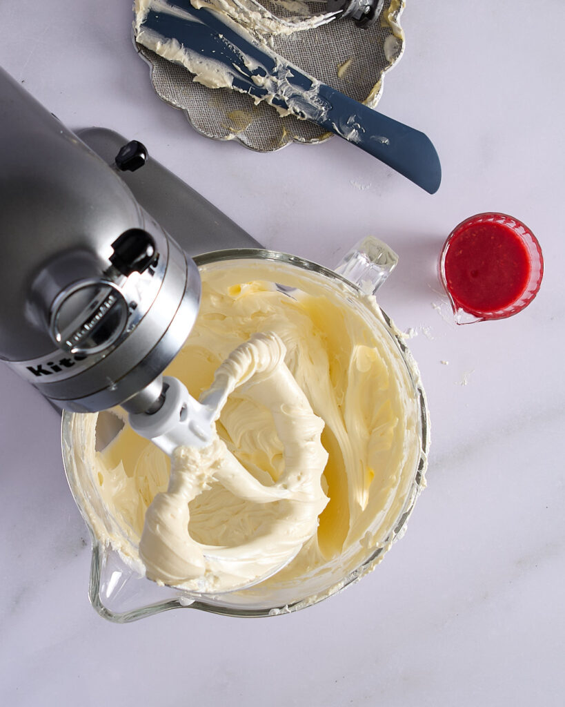 Butter fully incorporated into Swiss meringue, with raspberry purée in a jug ready to be mixed in to finish the raspberry Swiss meringue buttercream.