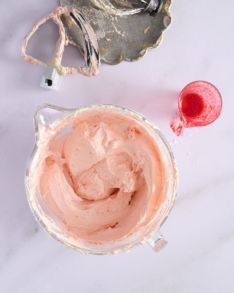 Bowl of finished raspberry Swiss meringue buttercream with a soft pink hue, next to a used paddle attachment and a nearly empty jug of raspberry purée.