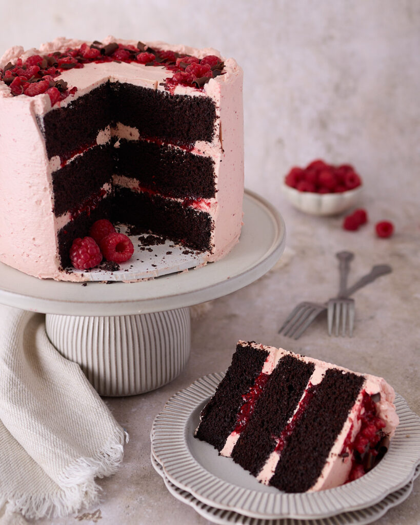 Three-layer chocolate raspberry celebration cake on a pedestal cake stand, with a slice removed and placed on a plate in the foreground. The cake is frosted with pale pink raspberry Swiss meringue buttercream, filled with raspberry jam, and topped with fresh raspberries and chocolate curls.