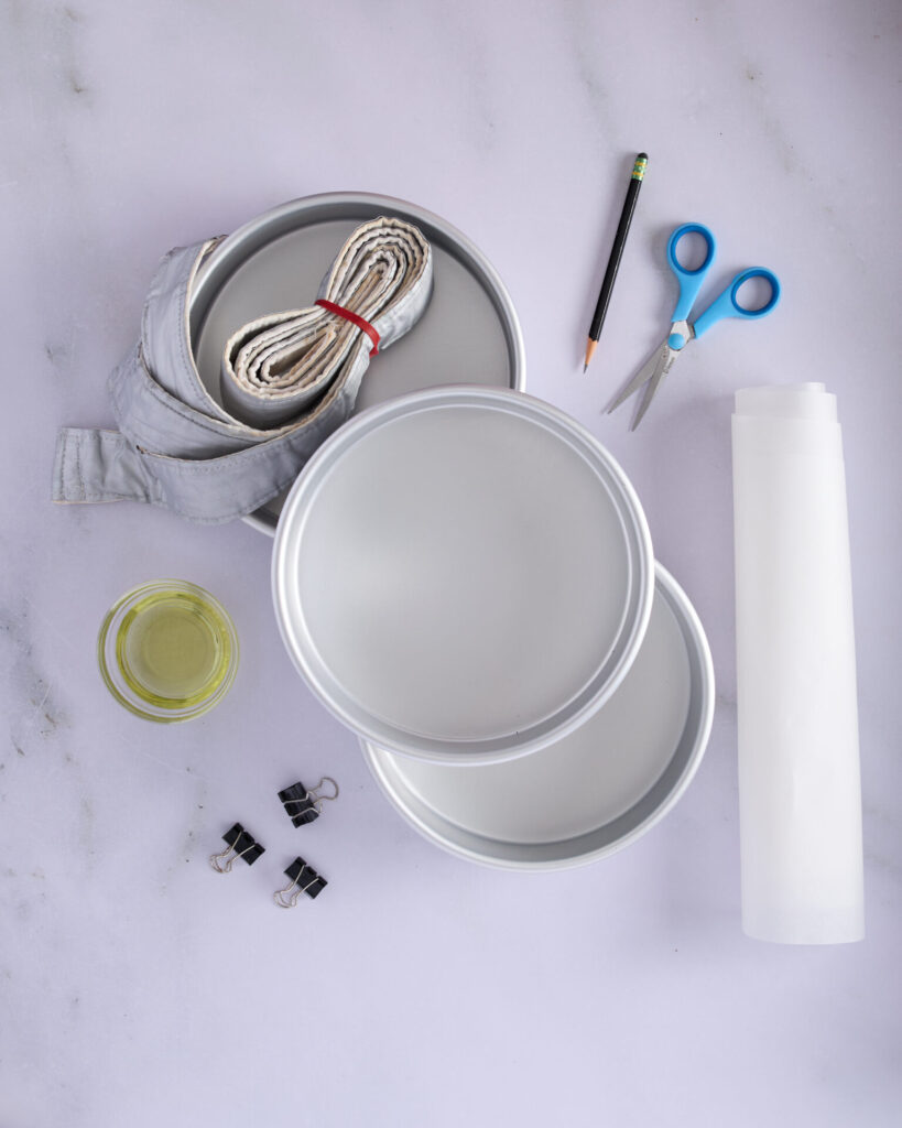 Cake prep tools for baking a three-layer chocolate cake, including cake pans, scissors, baking strips, baking paper, grapeseed oil, and bulldog clips.