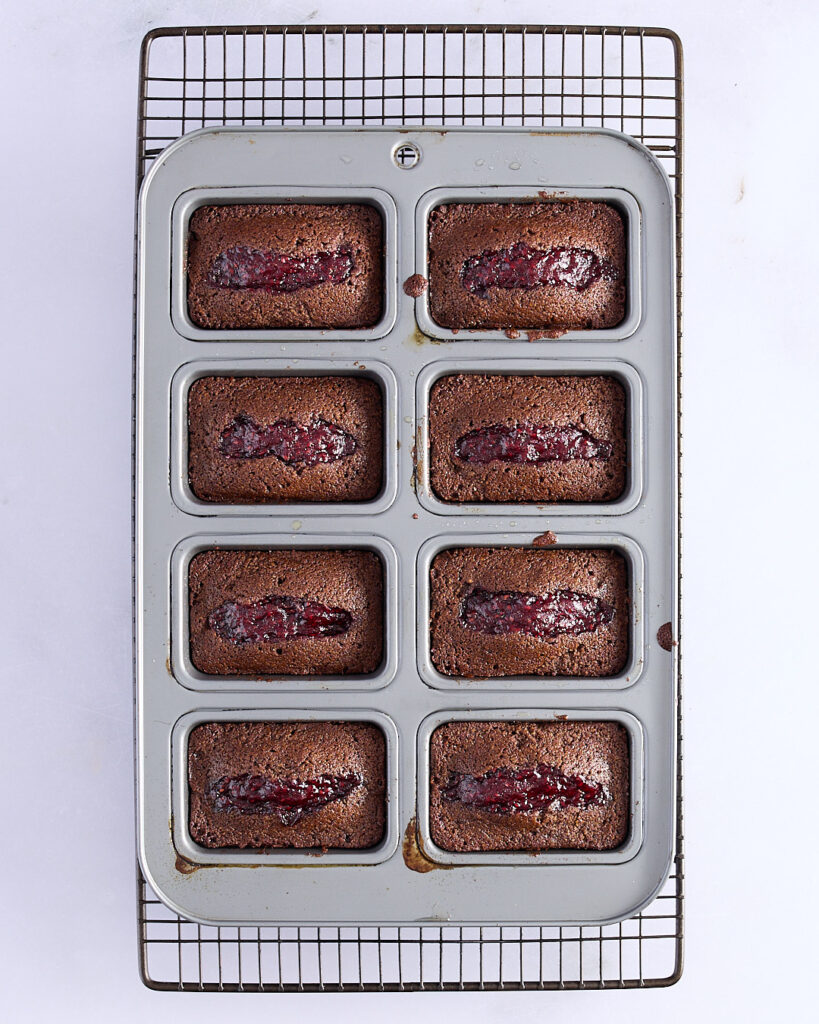 Freshly baked chocolate raspberry financiers in a mini loaf tin, resting on a cooling rack. Each cake has a visible raspberry jam centre.