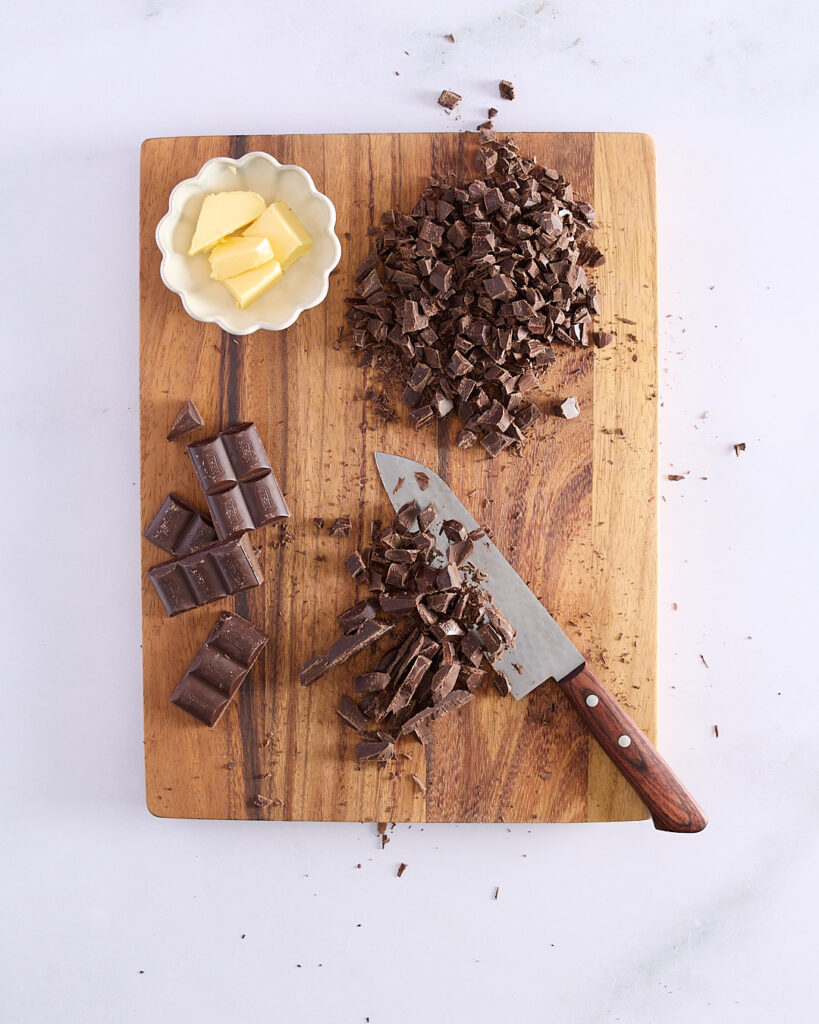 Chopped dark chocolate on a wooden board next to a knife and a small bowl of butter, ready for making ganache.