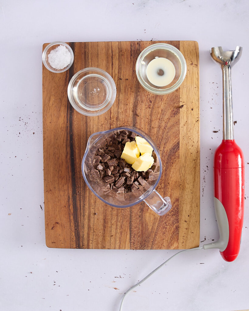 Chopped dark chocolate and butter in a jug surrounded by ingredients for ganache—salt, glucose, and cream—with an immersion blender alongside.