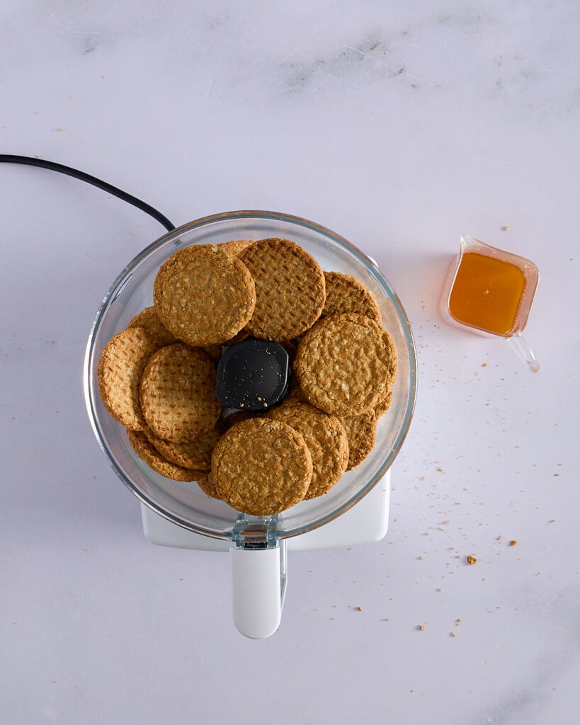 Whole sweet oat biscuits in a food processor next to a jug of melted brown butter, ready to be blitzed for a crumb crust.