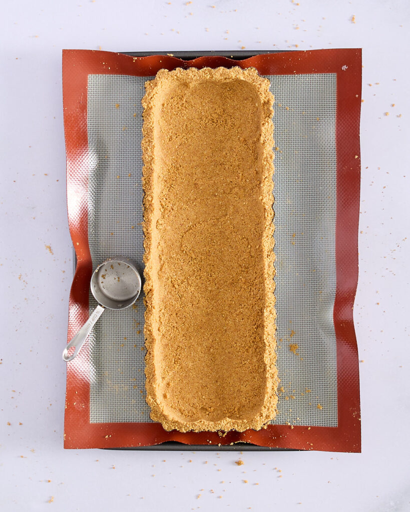 A rectangular tart tin filled with evenly pressed biscuit crumb base, sitting on a silicone mat with a measuring cup beside it.