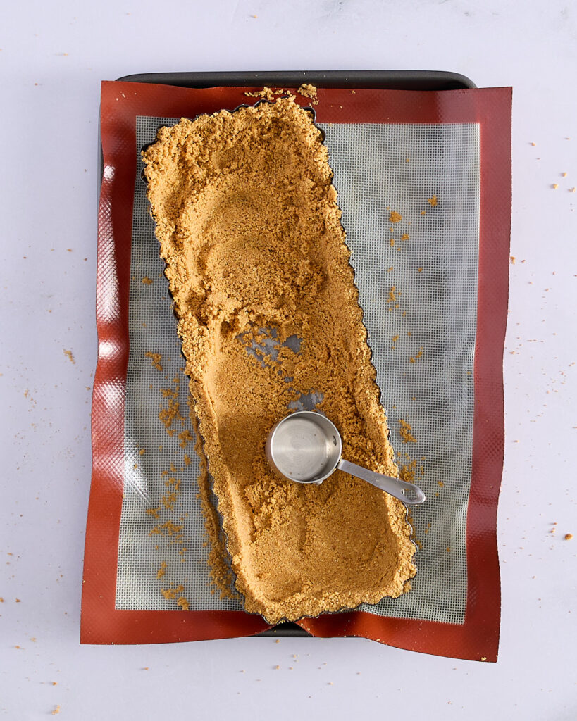 A tart tin halfway filled with biscuit crumbs, being pressed into shape with a metal measuring cup on a silicone baking mat.