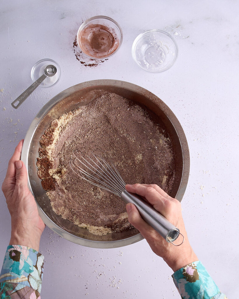 Sandra whisking dry ingredients in a mixing bowl, blending cacao powder with almond meal, sugar, cornflour, and salt.