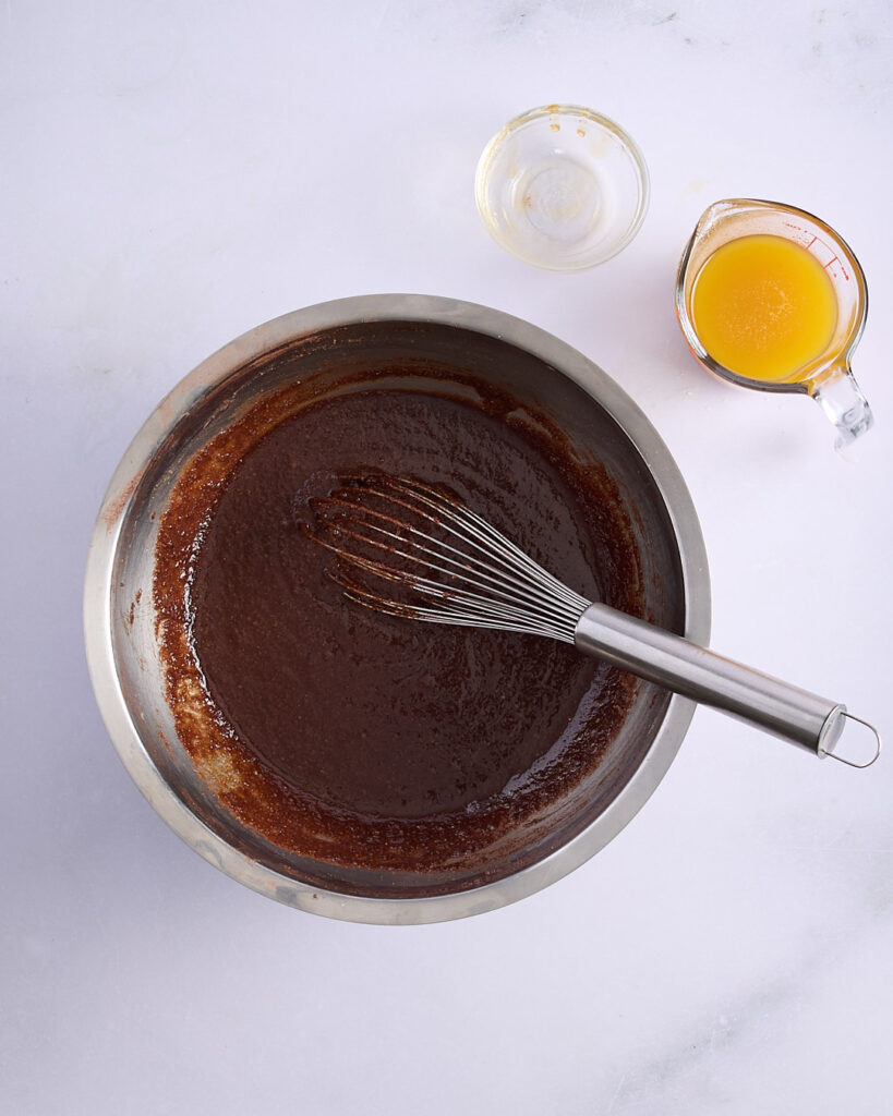 A mixing bowl of chocolate financier batter with a whisk, next to a jug of browned butter and a small bowl of honey on a white marble surface.