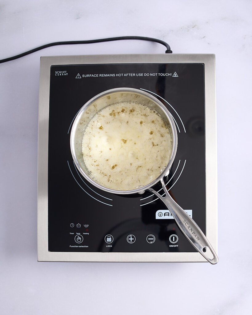Sandra stirring golden foaming butter with a silicone spatula on a cooktop, nearing the browned butter stage with milk solids visible.