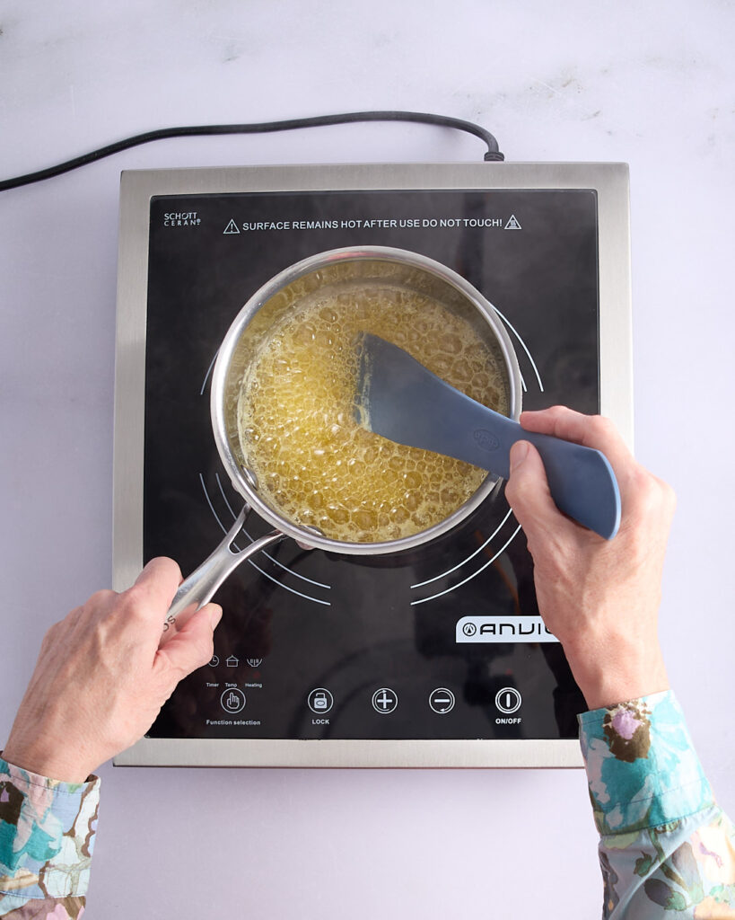 Sandra stirring golden foaming butter with a silicone spatula on a cooktop, nearing the browned butter stage with milk solids visible.