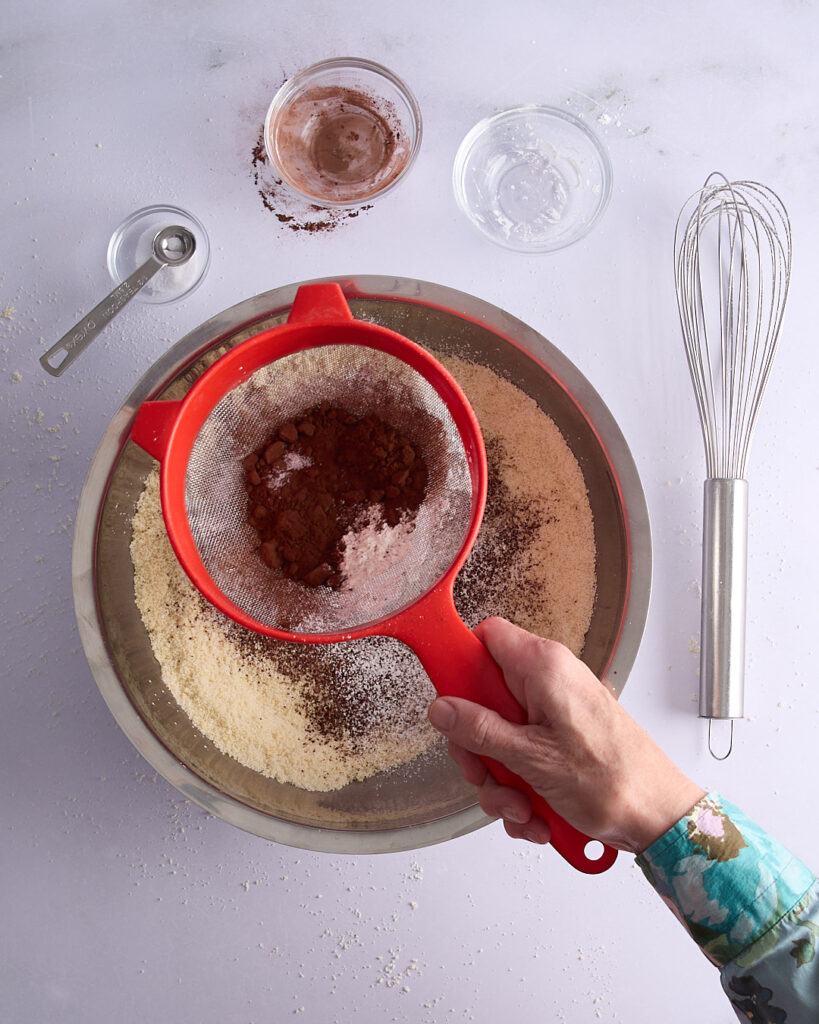 Sandra sifting cacao powder and dry ingredients into a large metal mixing bowl of almond meal and sugar, with a whisk and measuring spoons nearby.
