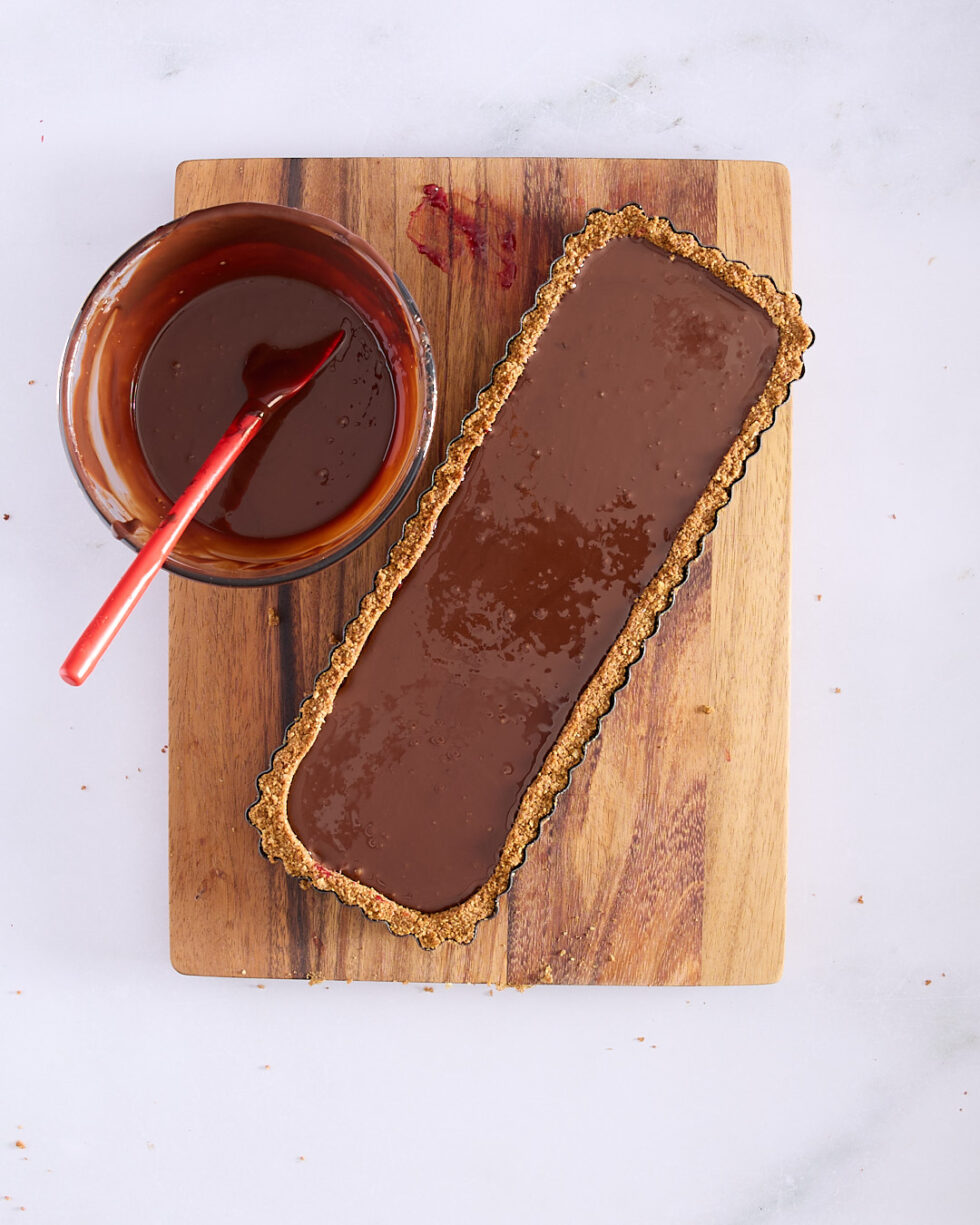 A rectangular tart shell filled with glossy dark chocolate ganache, sitting on a wooden board next to a bowl of leftover ganache with a red spatula.