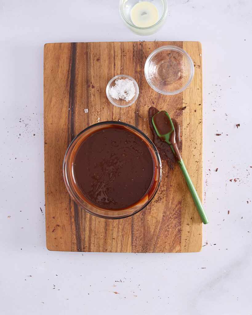 A bowl of freshly blended dark chocolate ganache with a green spatula, surrounded by leftover ingredients on a wooden board.