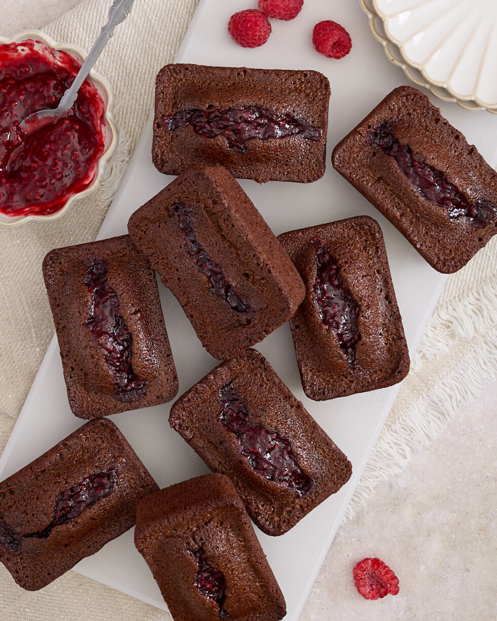 A tray of chocolate raspberry financiers, each topped with a swirl of raspberry jam, surrounded by fresh raspberries and a bowl of jam on a cream cloth.