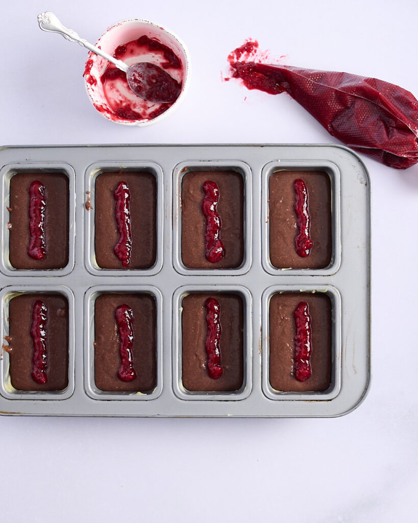 Low sugar raspberry jam piped neatly onto of each portion of chocolate financier batter,with a piping bag of jam and empty dish of jam with a spoon resting in it.