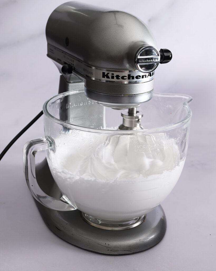 A stand mixer whipping glossy white marshmallow topping in a glass bowl, mid-process for the s’mores tart recipe.